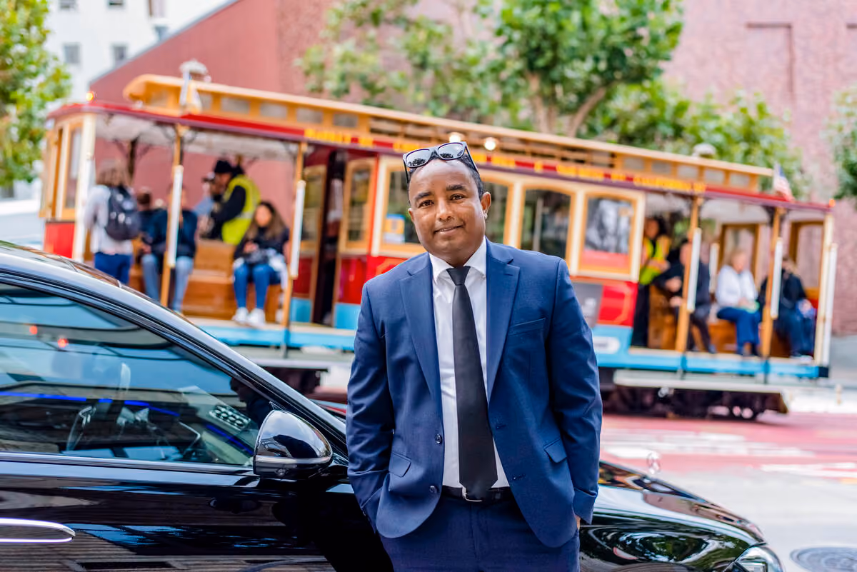 Chauffeur standing in front of a black car as a cable car full of passengers passes behind