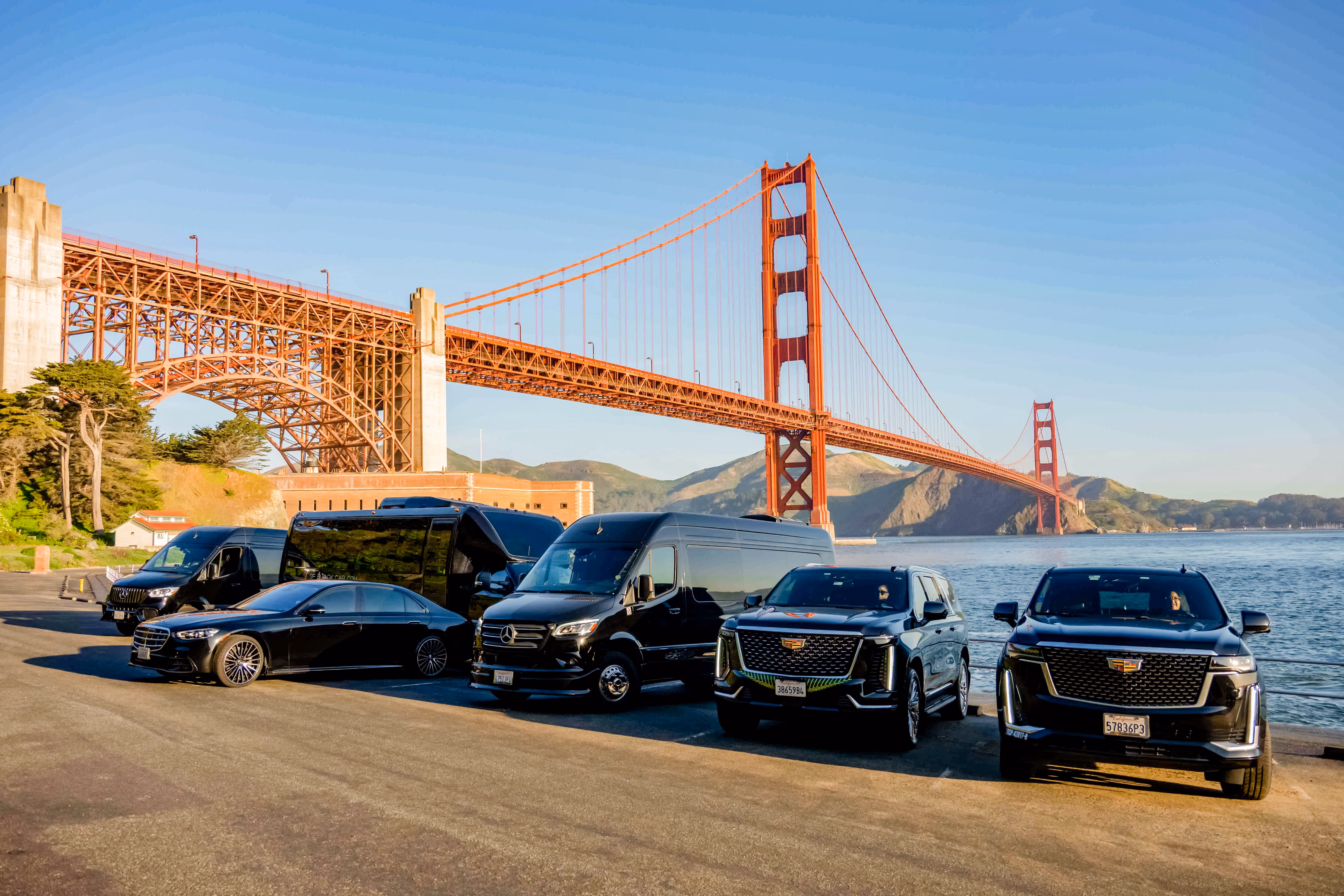 Fleet of black vans, a sedan and two Cadillac SUVs parked below a red suspension bridge