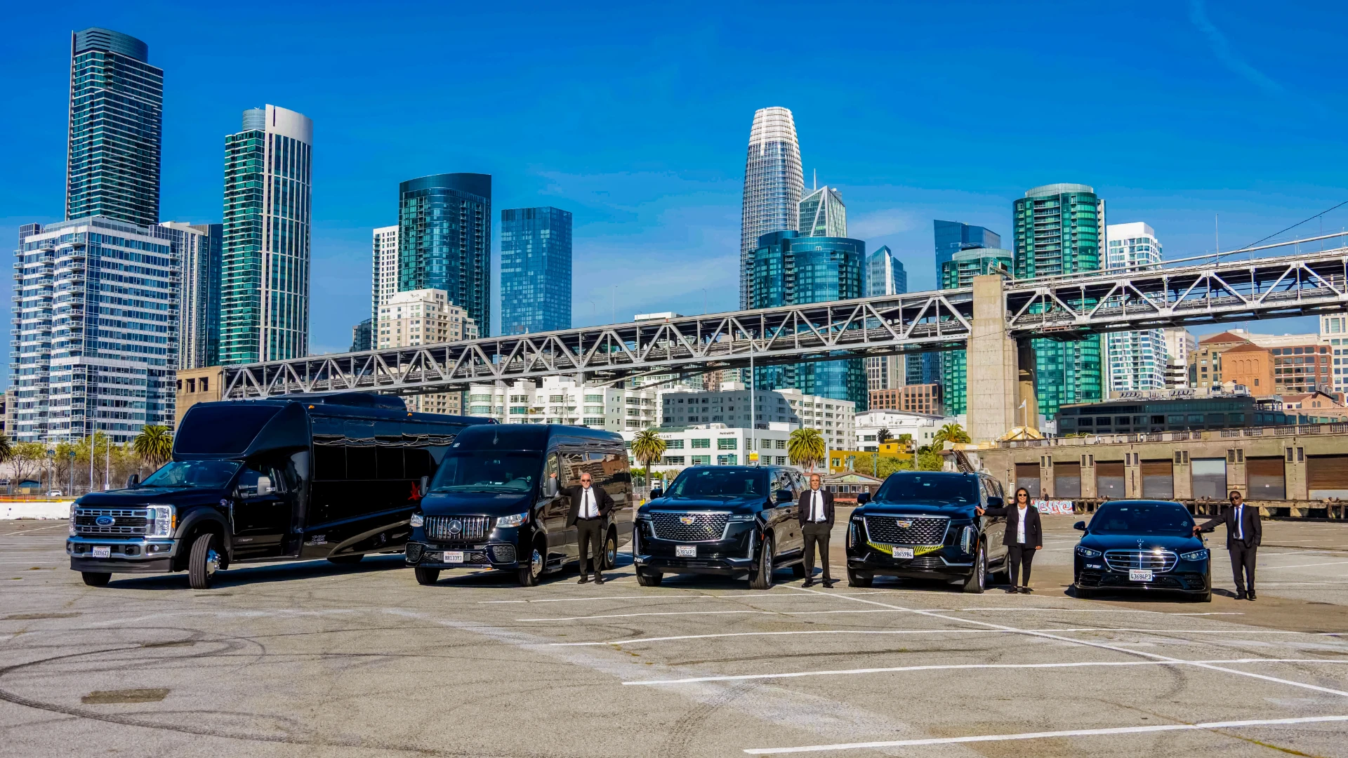 Chauffeurs standing with a fleet of black vehicles in a wide lot with skyscrapers and a bridge behind