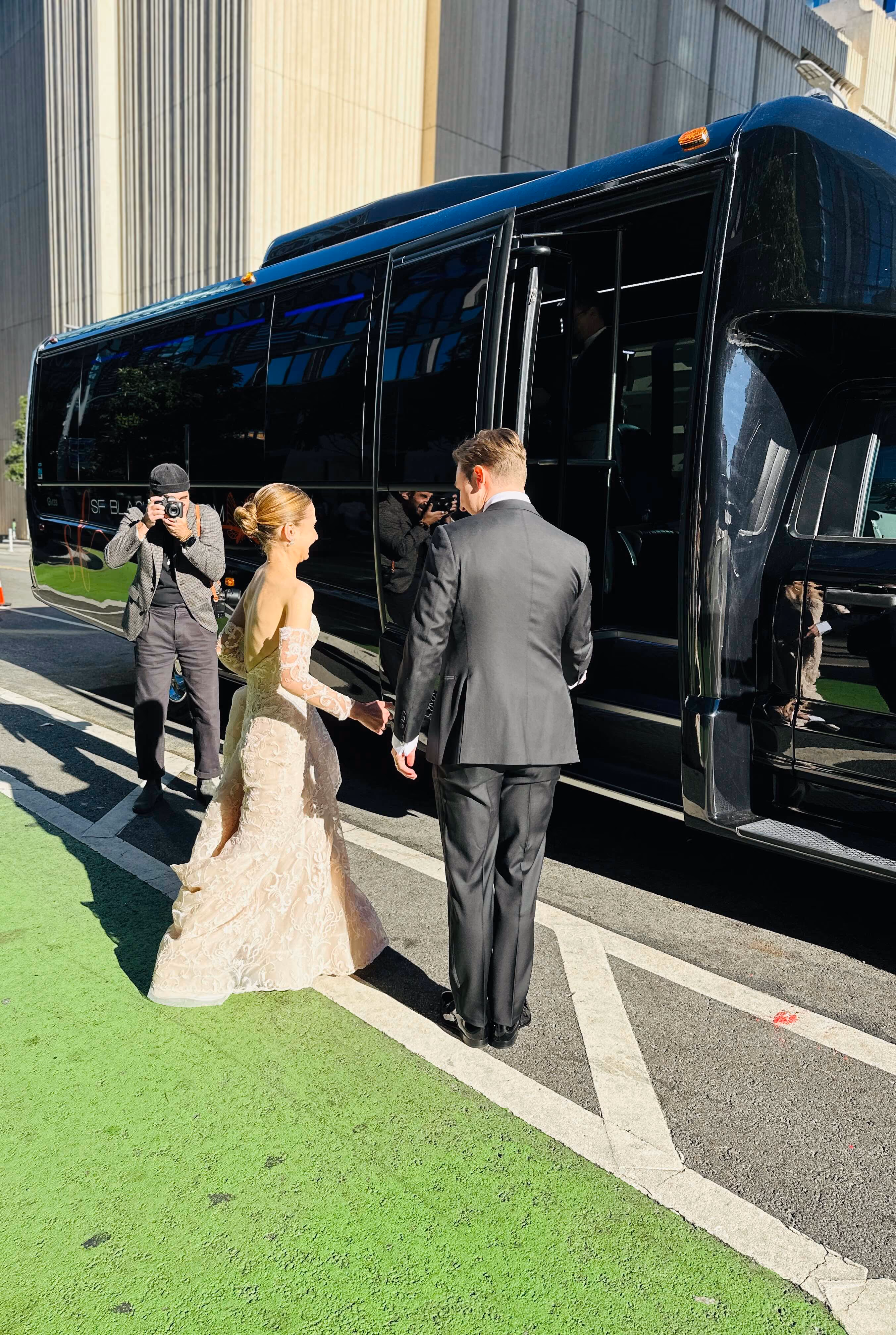 Bride and groom walking toward a black limo bus while a photographer takes pictures