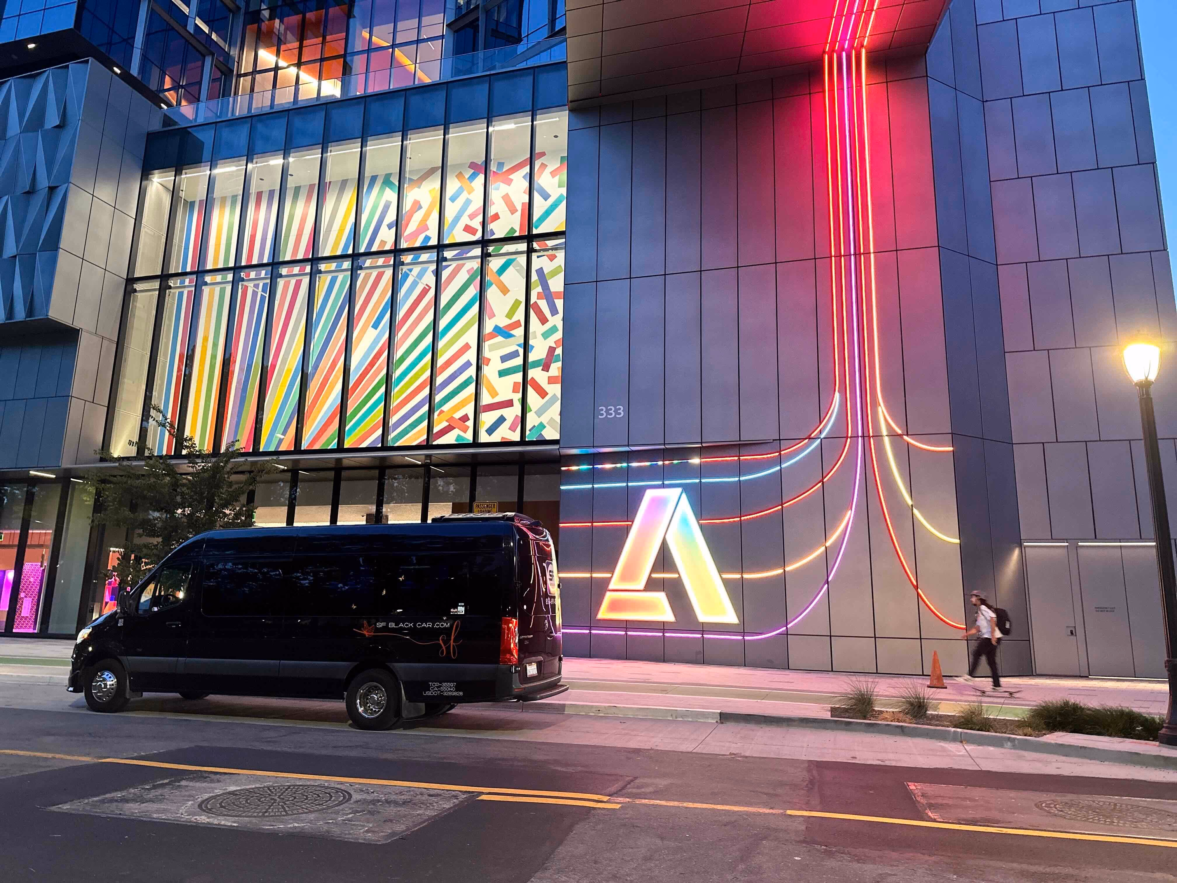 Black SF Black Car Sprinter van parked on a street outside an illuminated modern glass building at dusk
