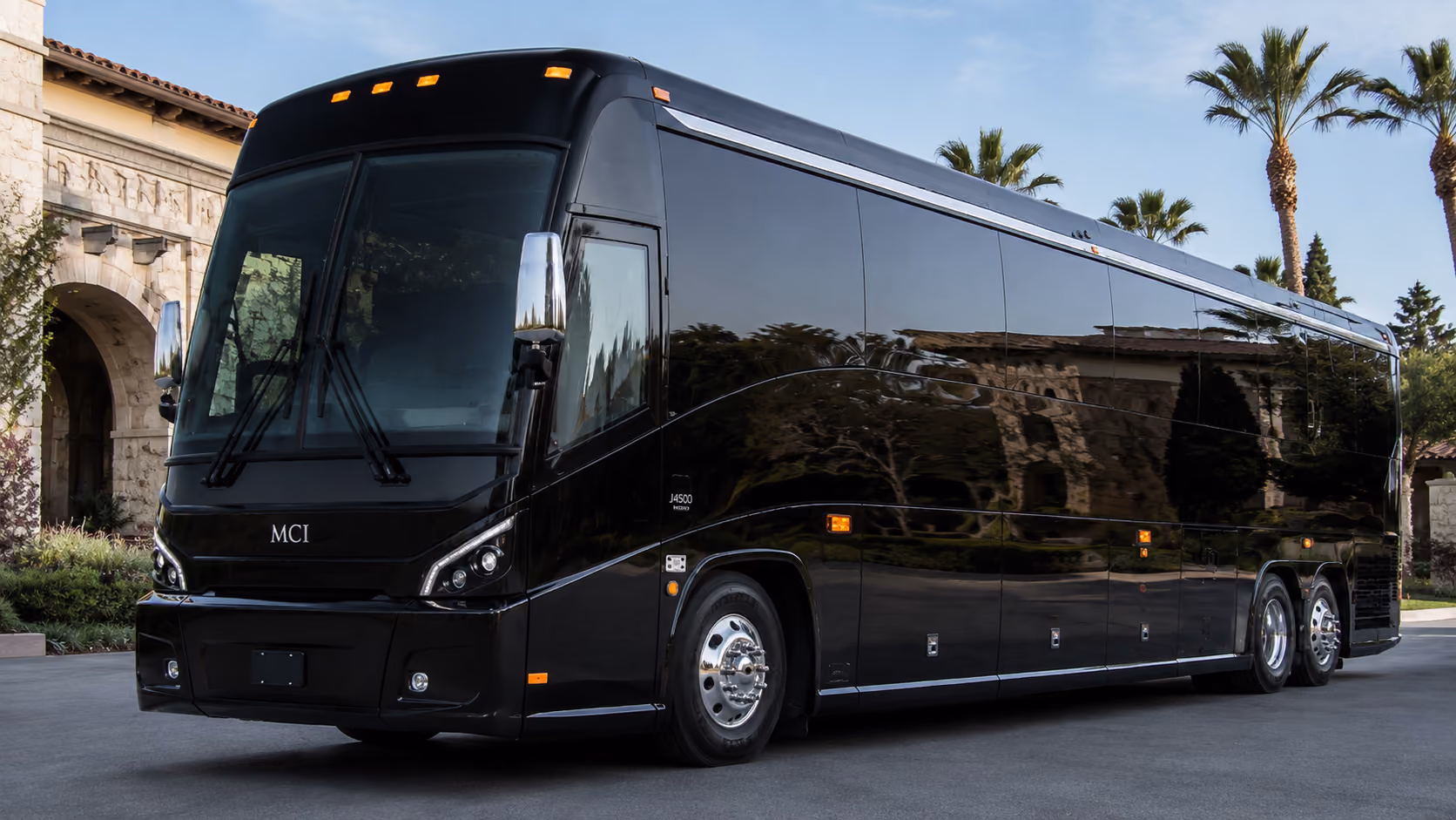 Shiny black motor coach bus parked with palm trees and a stone building in the background.