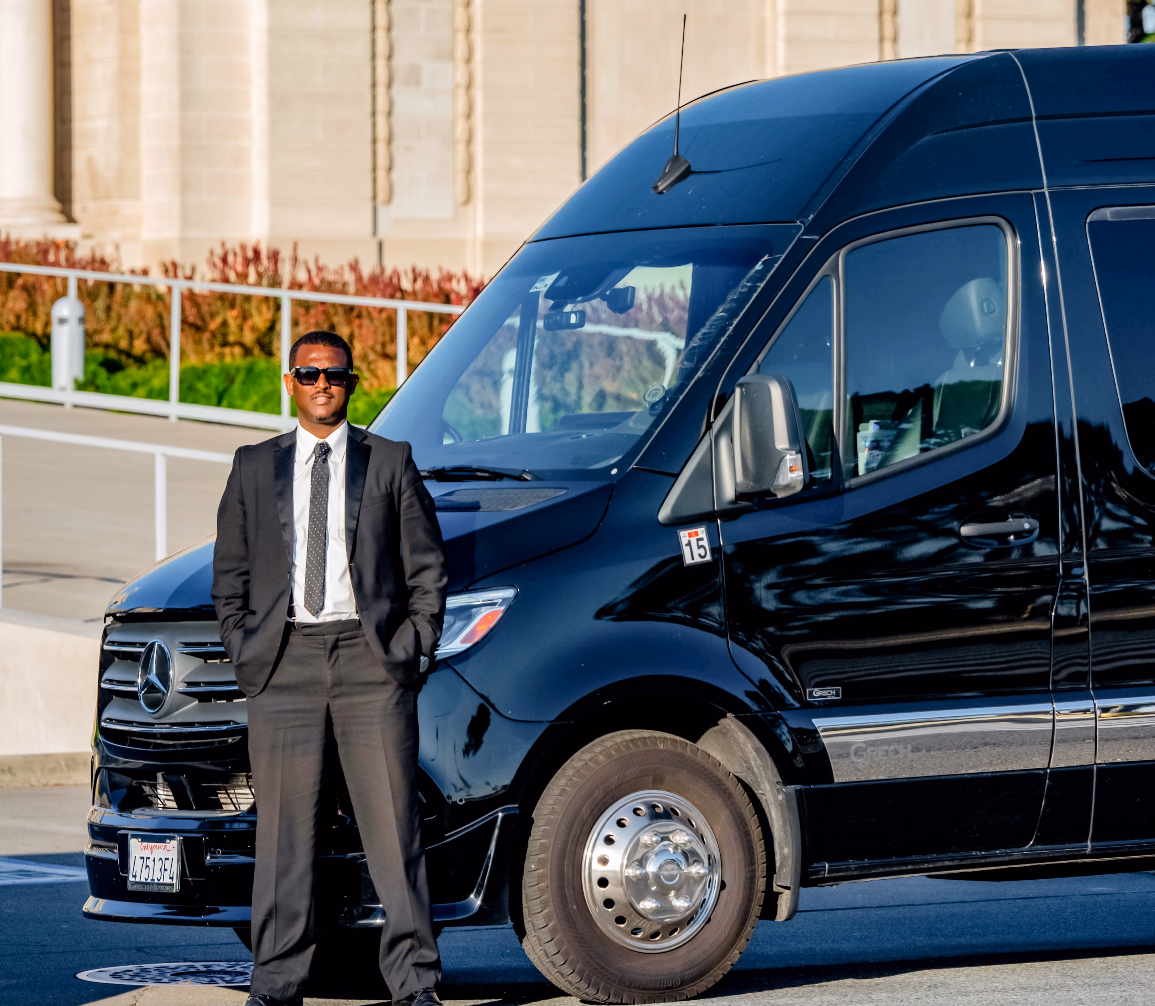 Chauffeur in a dark suit and sunglasses standing beside a black Mercedes Sprinter van