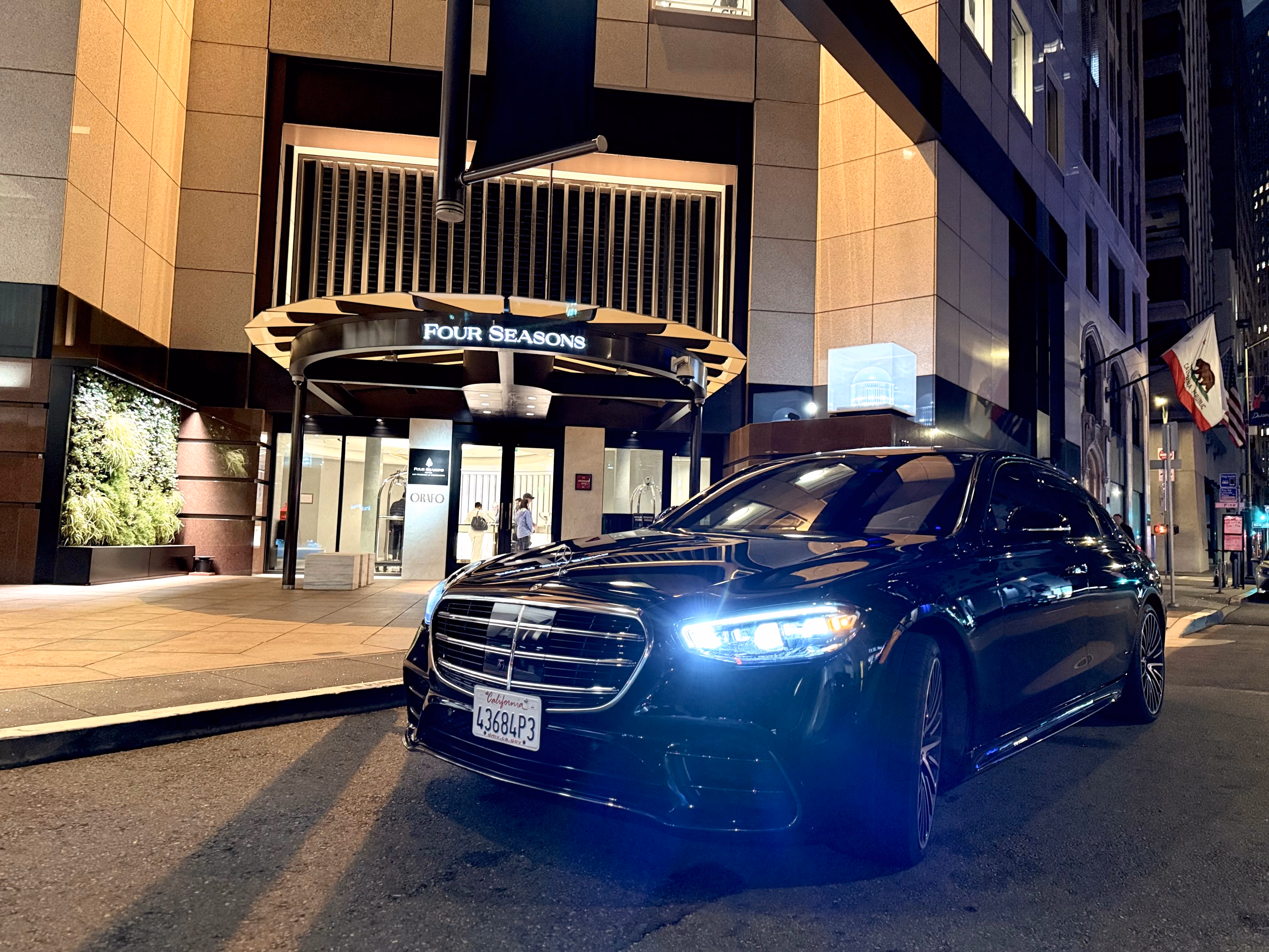 Black Mercedes sedan with headlights on at the lit Four Seasons hotel entrance at night
