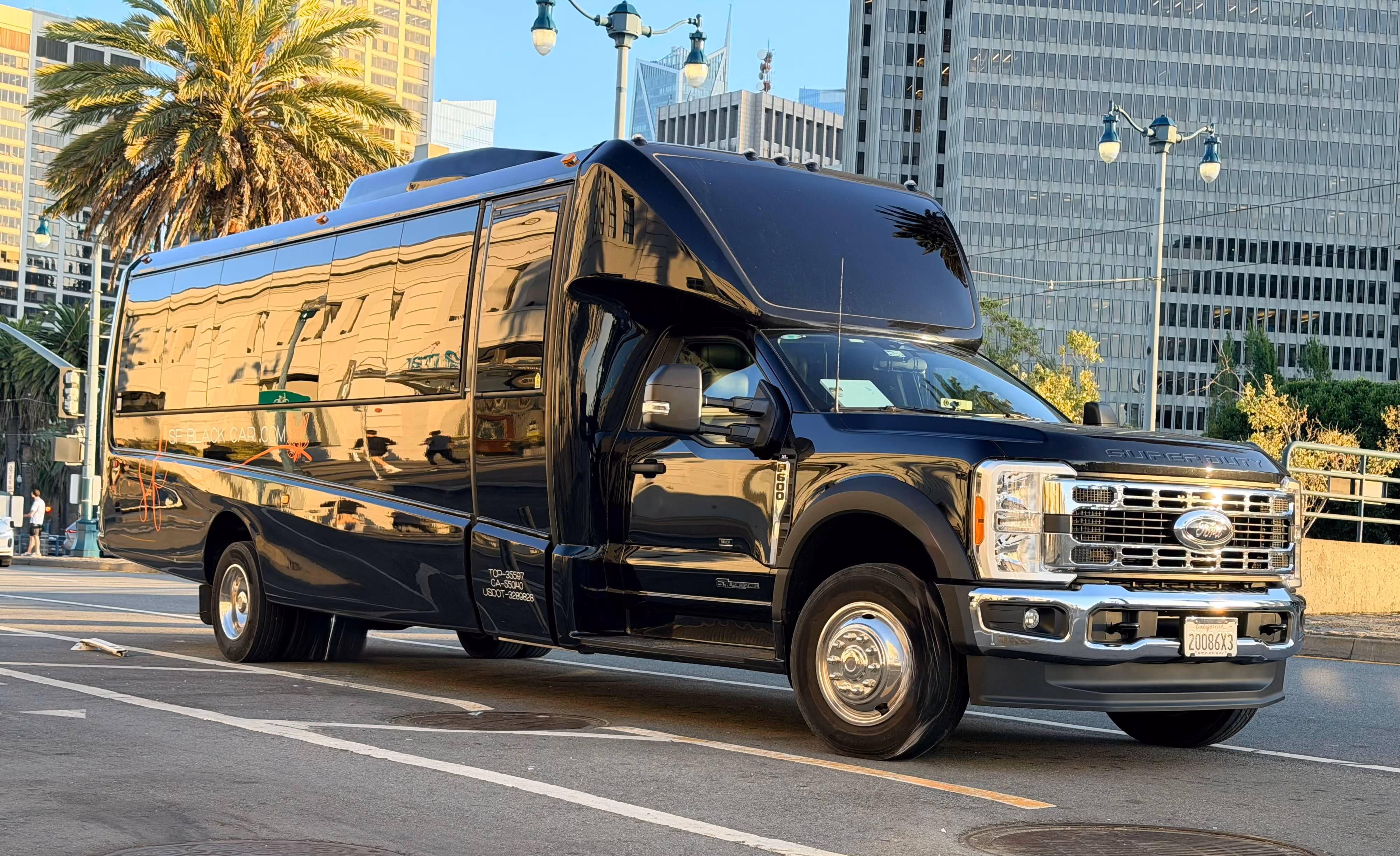 Black Ford Super Duty shuttle bus parked on city street with palm trees and buildings nearby.