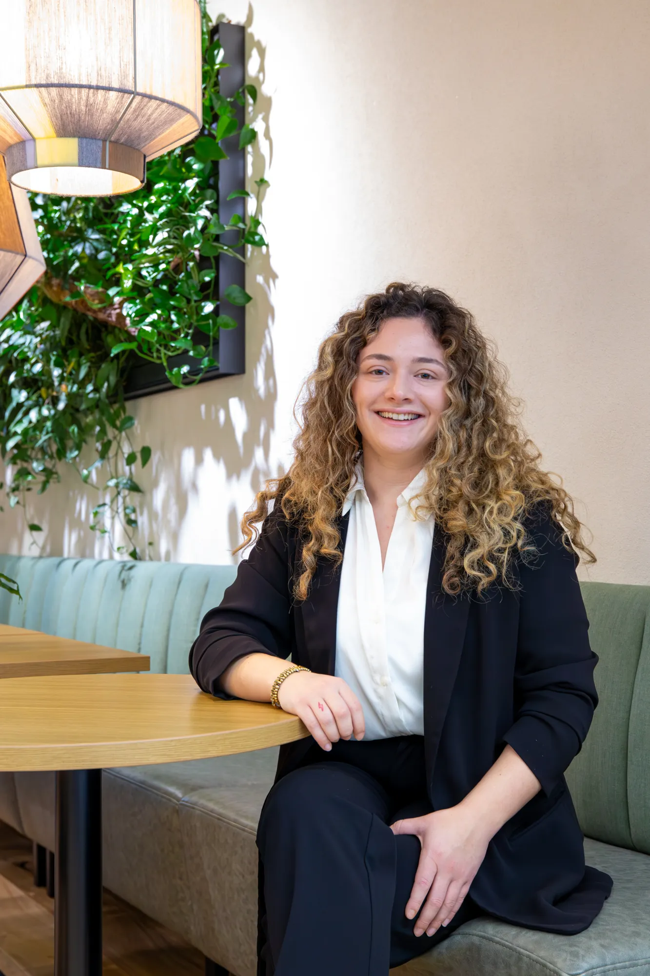 Smiling woman with curly hair wearing a black blazer and white shirt sitting on a green cushioned bench at a wooden table in a well-lit room with hanging lamps and green plants on the wall.