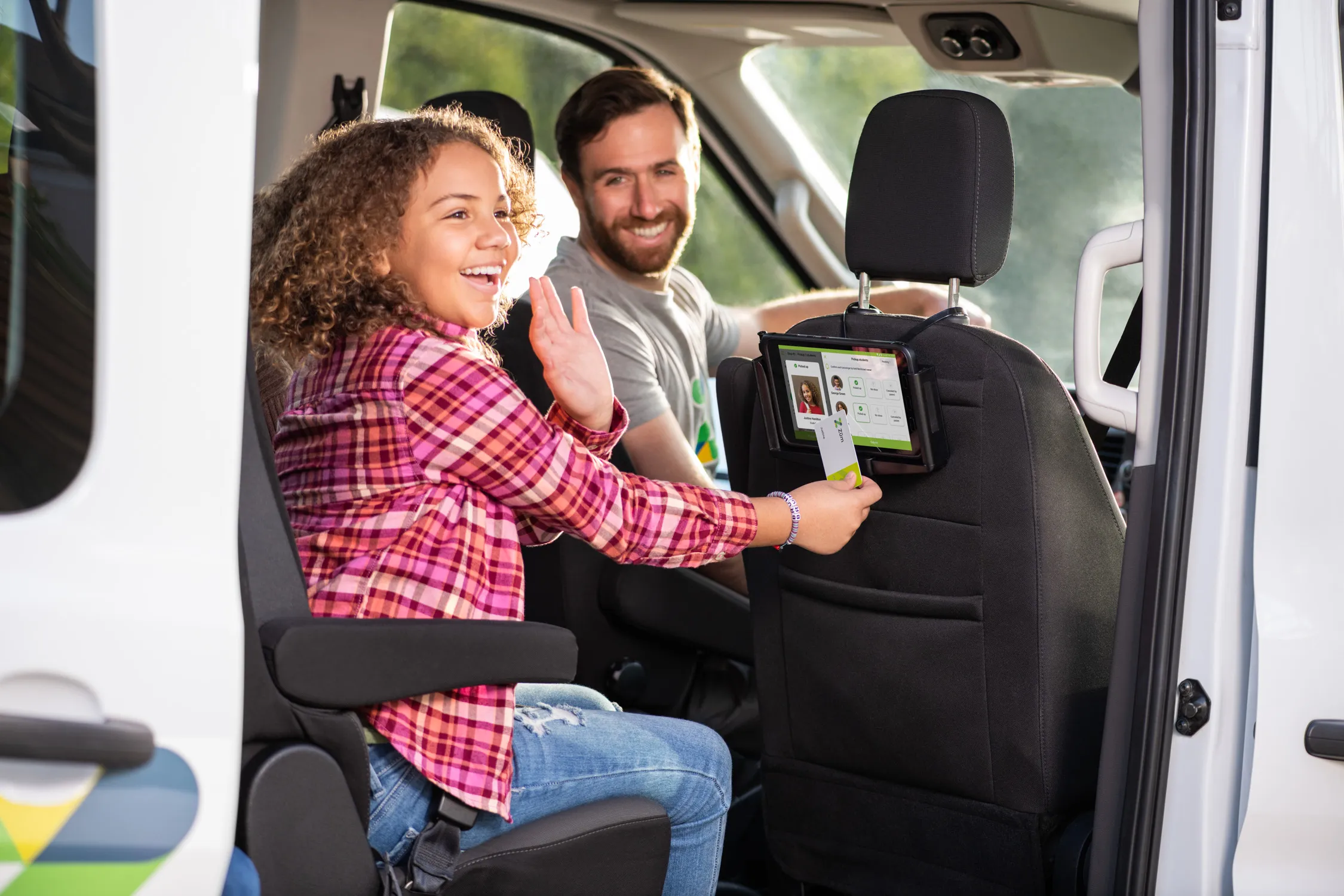 Child and driver smiling in a van, with a tablet mounted on the seat in front of them.