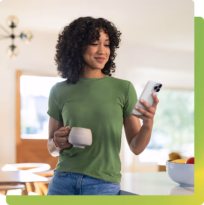 Woman with curly hair holding a cup and smartphone, smiling while standing in a modern kitchen.