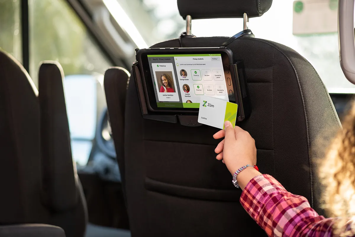 A child scans a card on a tablet mounted to a bus seat, showing a user interface.