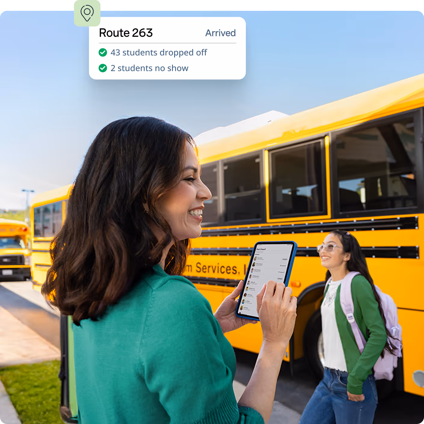A woman smiles while using a tablet near yellow school buses, with a student walking by.