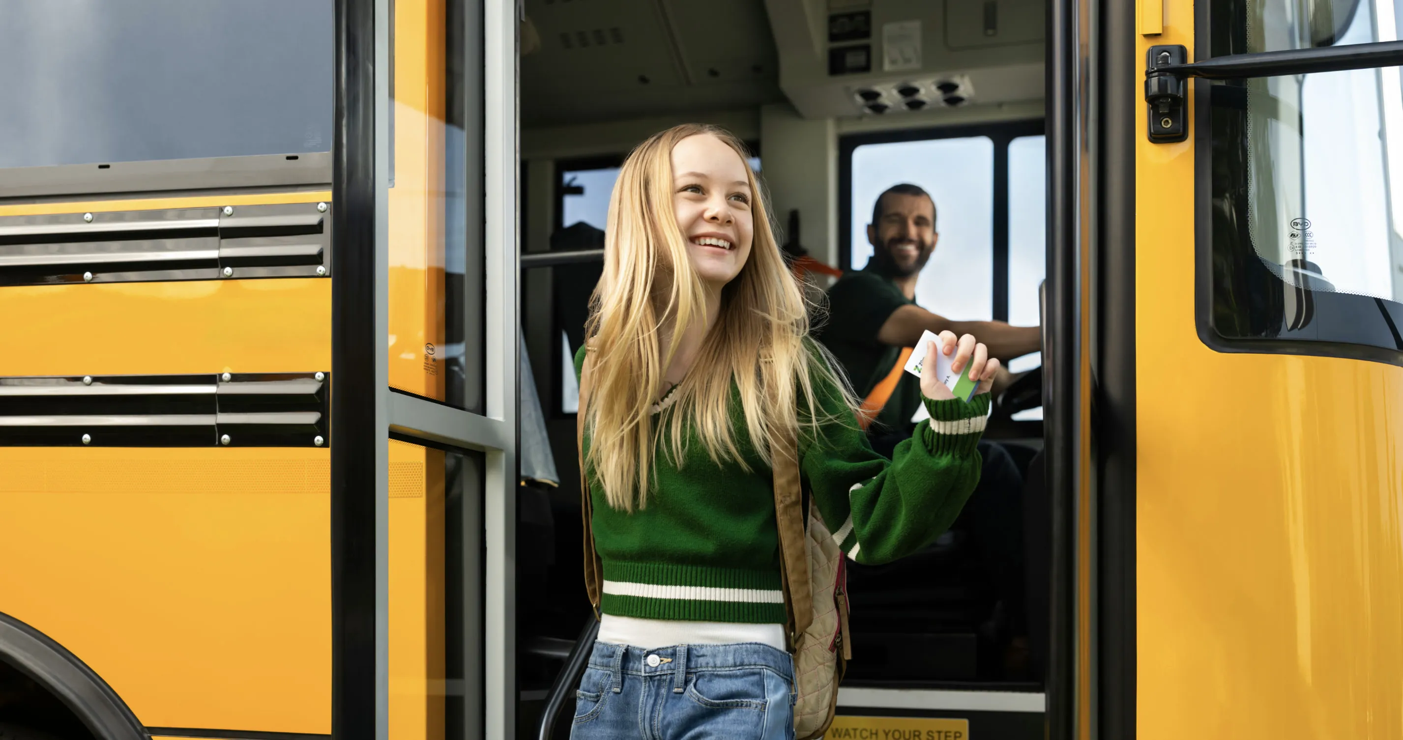 A girl with long hair smiles as she exits a school bus, with a driver visible in the front.