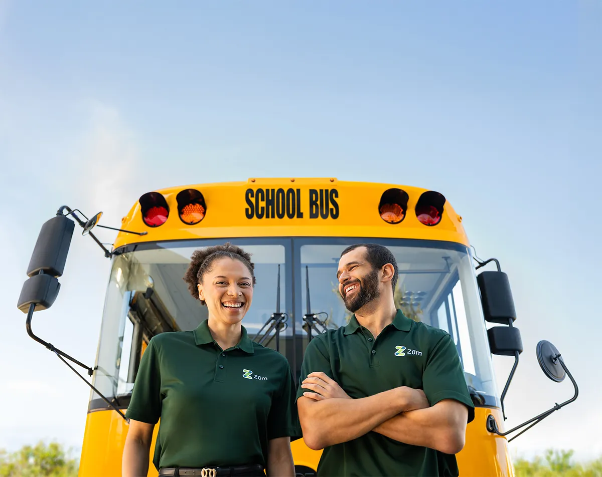 Two smiling Zum Services, Inc. drivers standing in front of a school bus.