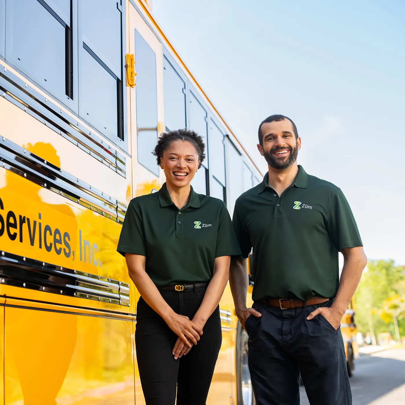 Two smiling employees of Zum stand beside a bright yellow school bus, wearing company polo shirts.