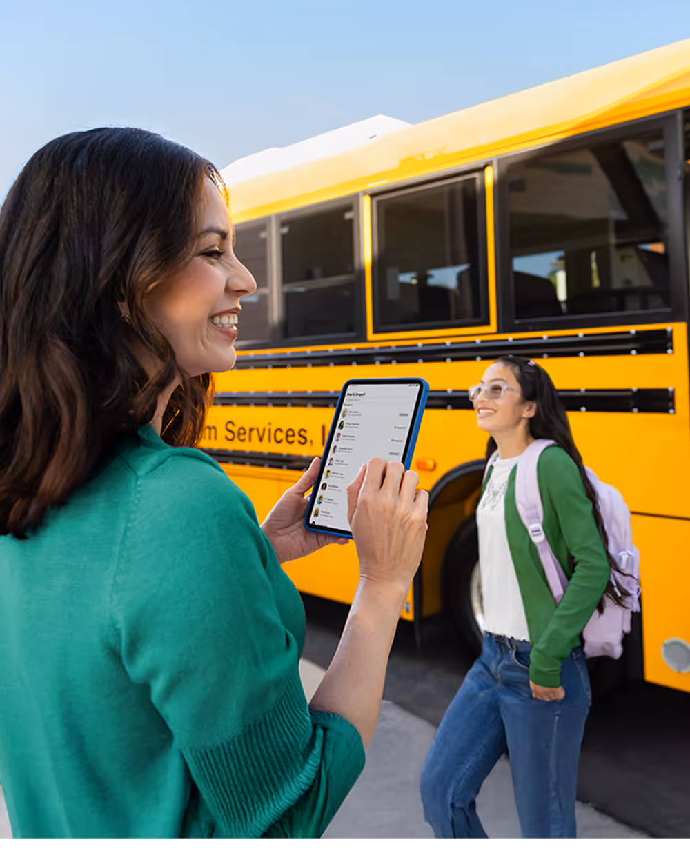 A staff member checking student pickup information on a tablet while a student walks toward a Zum Services, Inc. school bus.