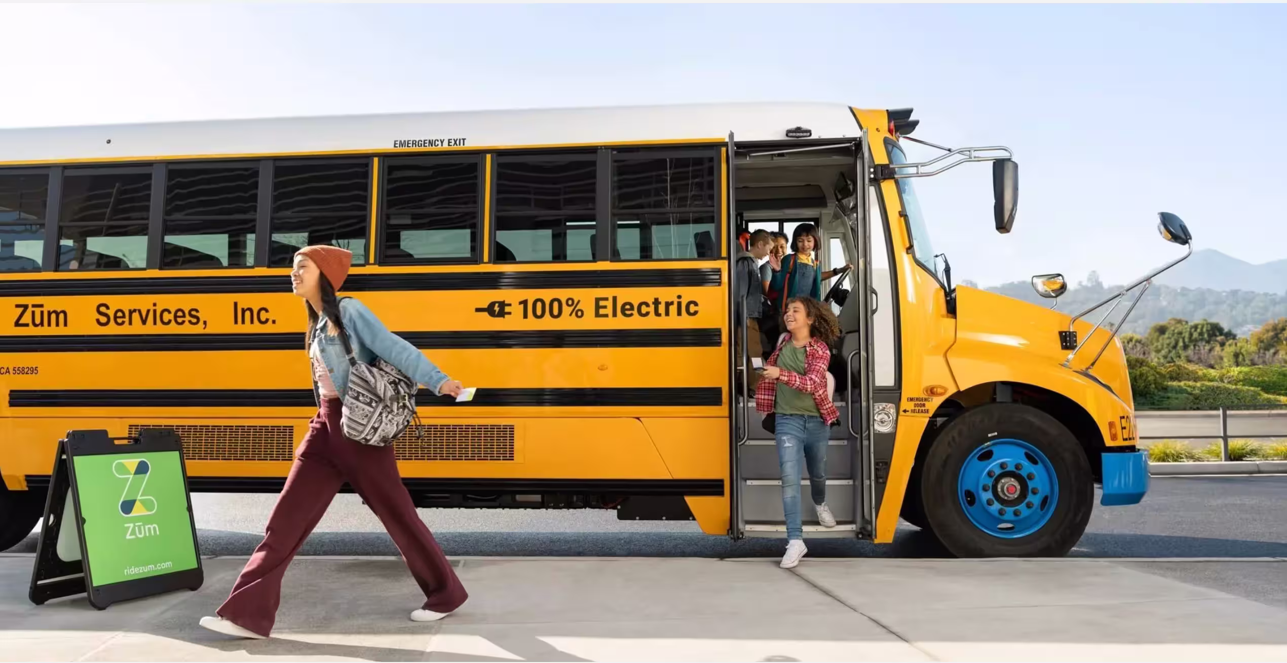 Students exiting a yellow electric bus while a girl runs towards it. Zum branding visible.