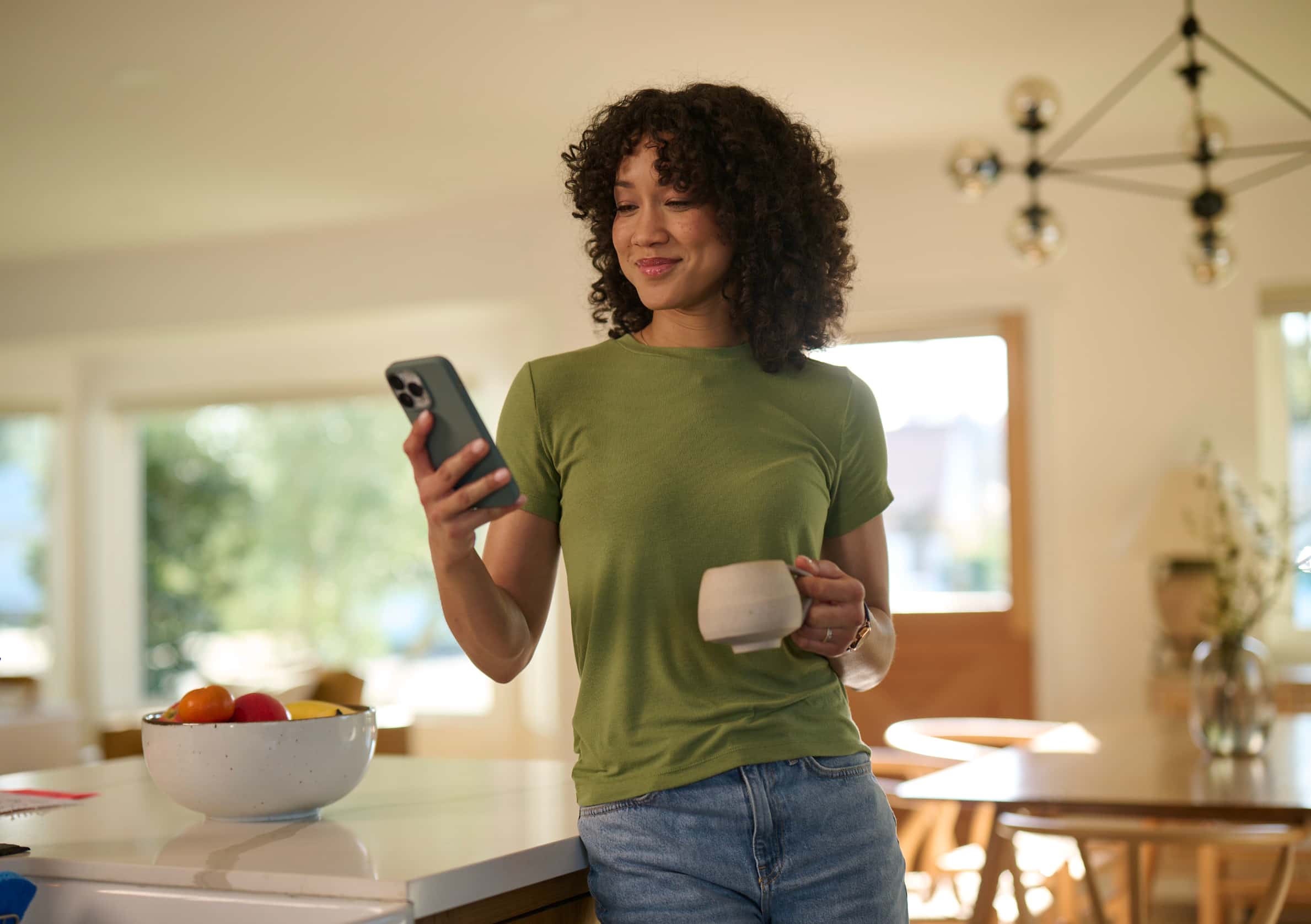 Woman with curly hair holding a cup and smartphone, smiling while standing near a kitchen table.