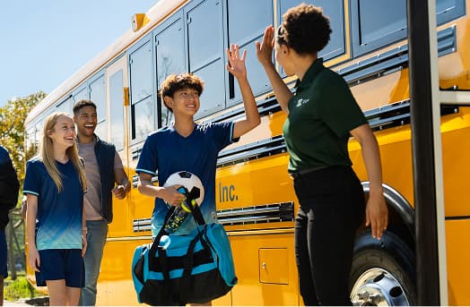 Students happily greet a driver while boarding a yellow school bus.