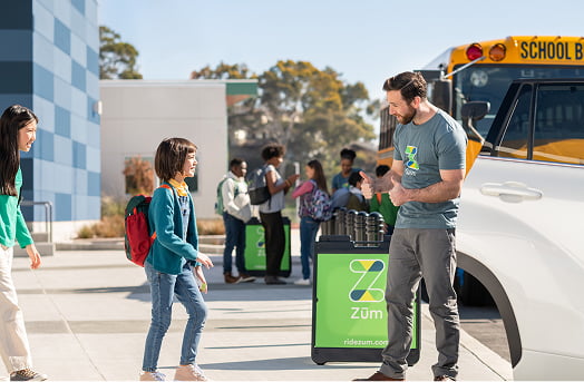 A driver assists a student with a backpack as other children gather near a school bus.