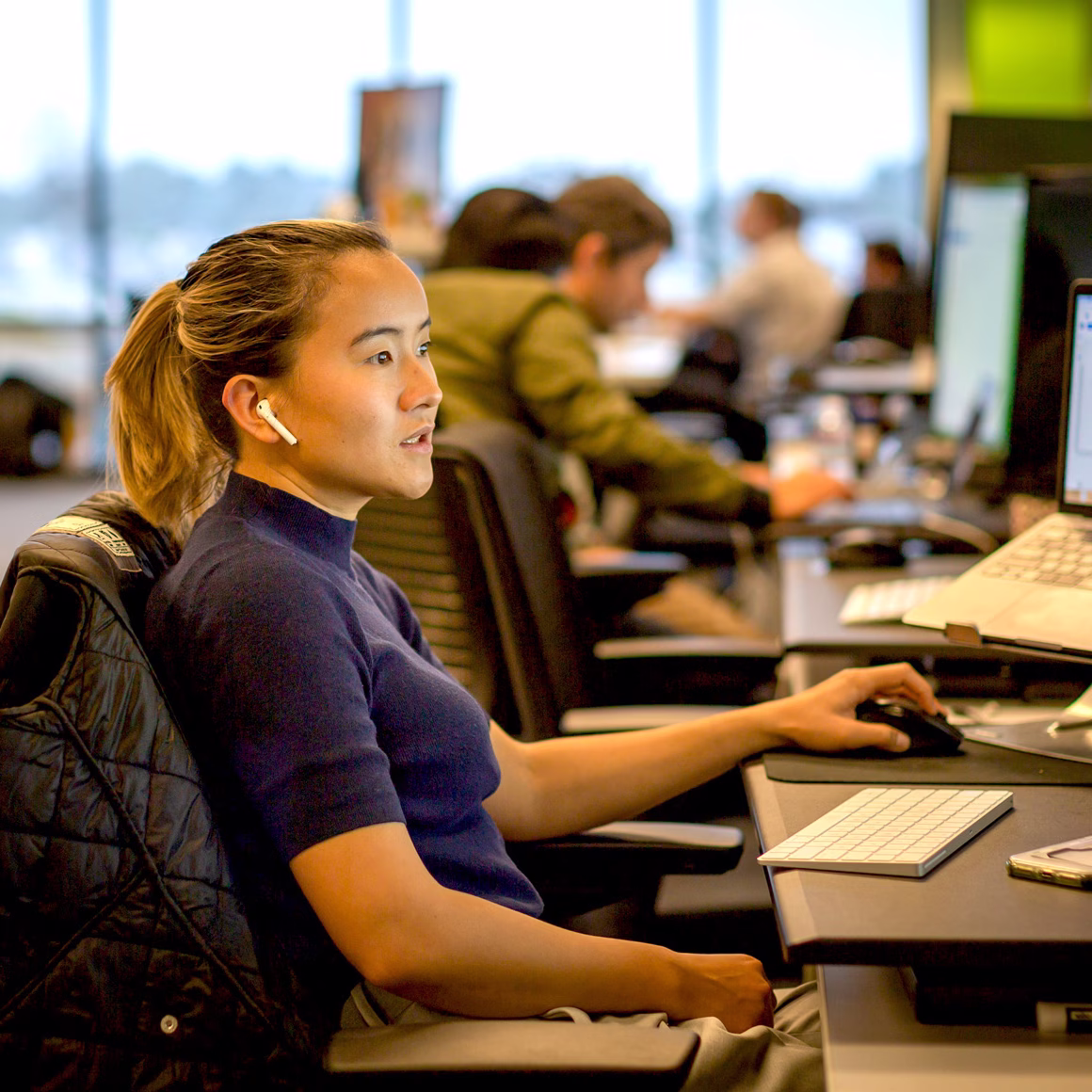 A Zum Services, Inc. employee wearing an earbud working at a desktop computer in an open office, with coworkers seated at desks in the background.