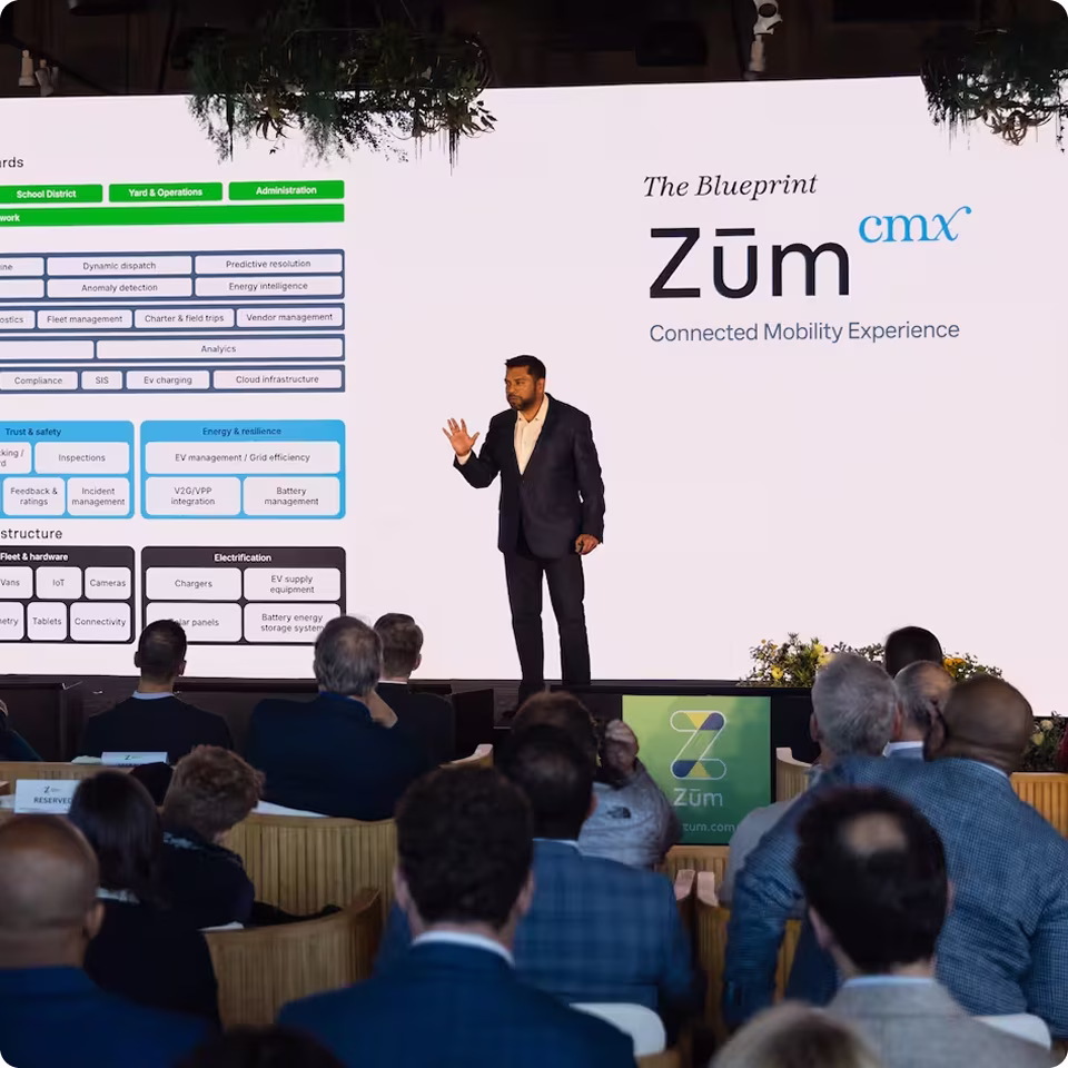 Speaker presenting on stage with a screen showing Zum CMX platform overview and audience in