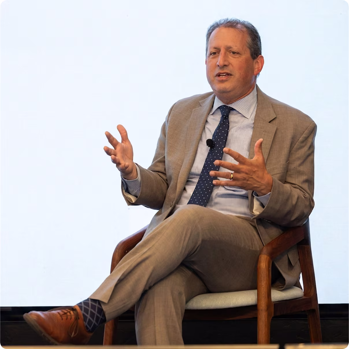Man in a suit sitting and gesturing, speaking during a presentation or discussion.
