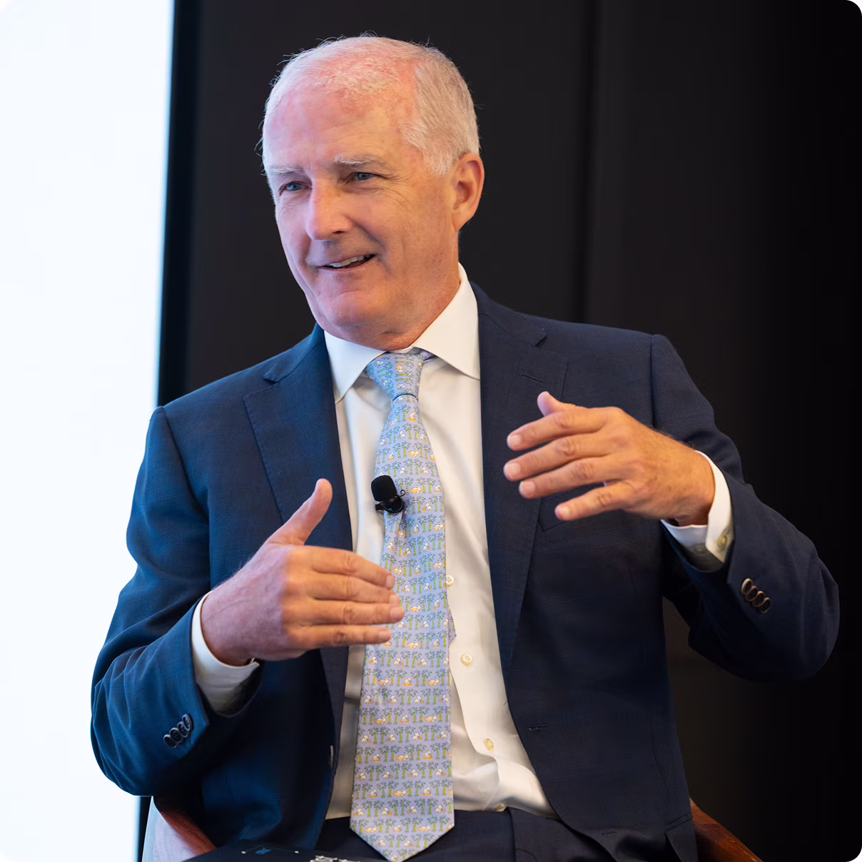 Man in a suit speaking at an event, gesturing with hands, seated in front of a screen.