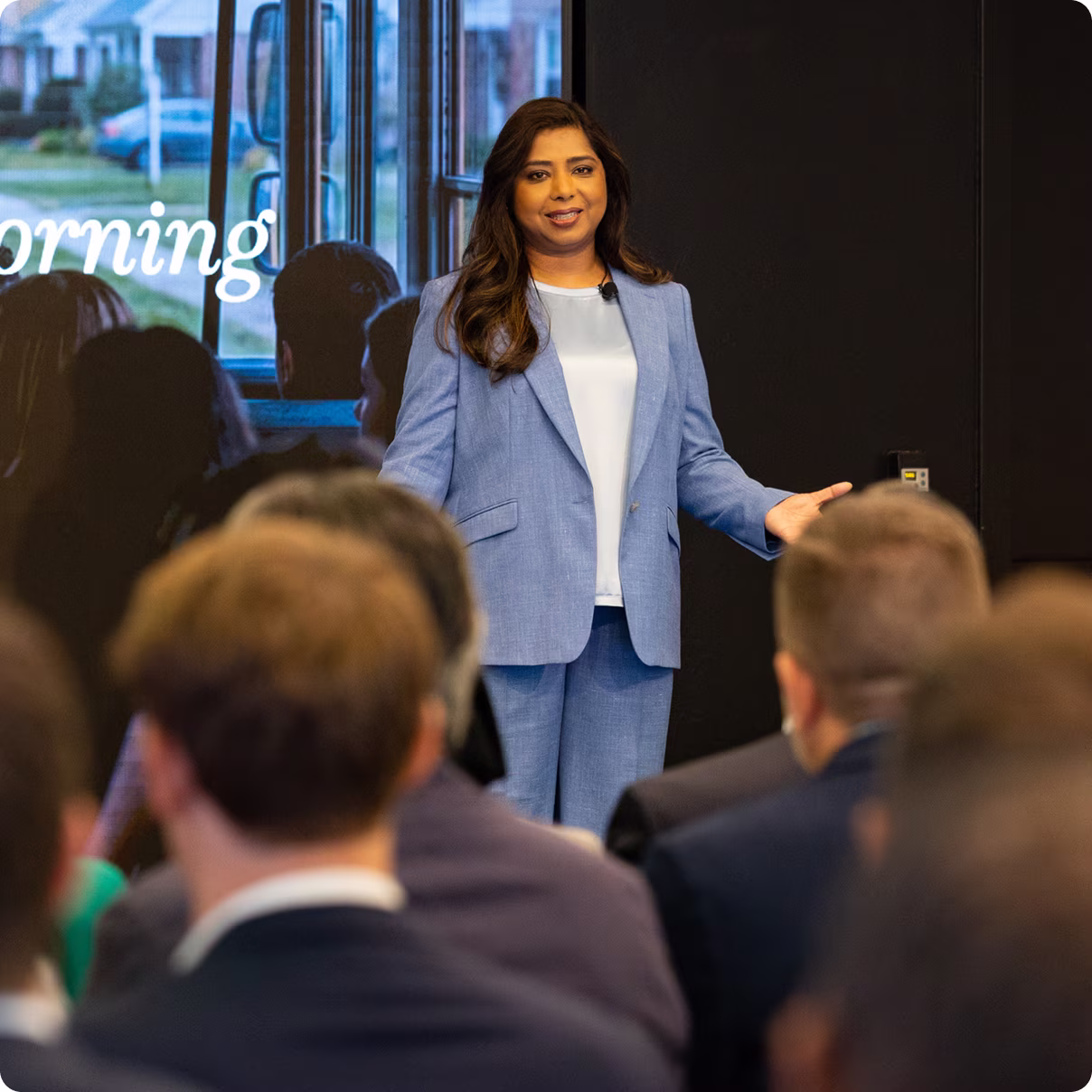 A speaker in a blue suit addresses an audience seated in front of her.