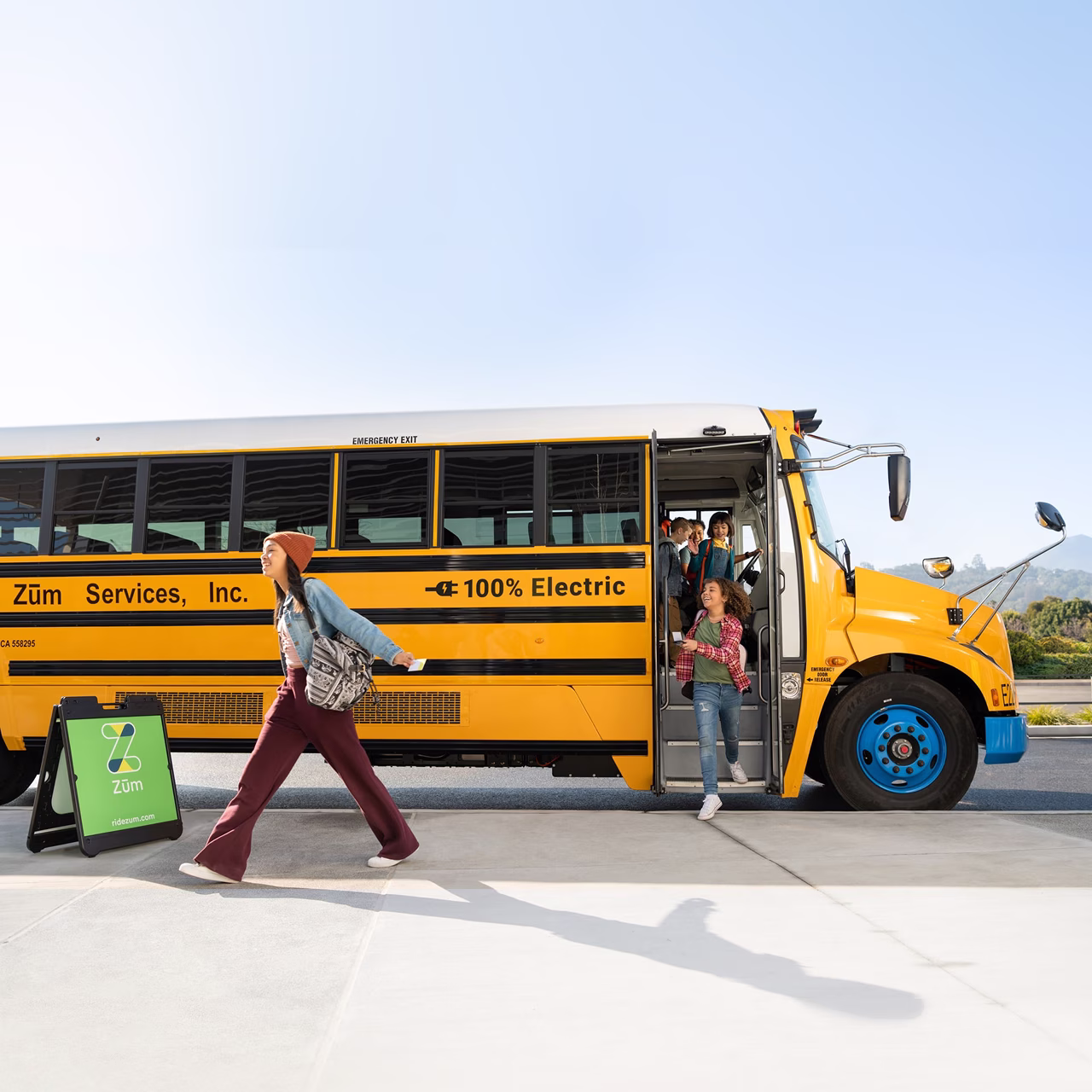 Students exiting a 100% electric Zum Services, Inc. school bus, with a Zum sidewalk sign displayed in front of the vehicle.