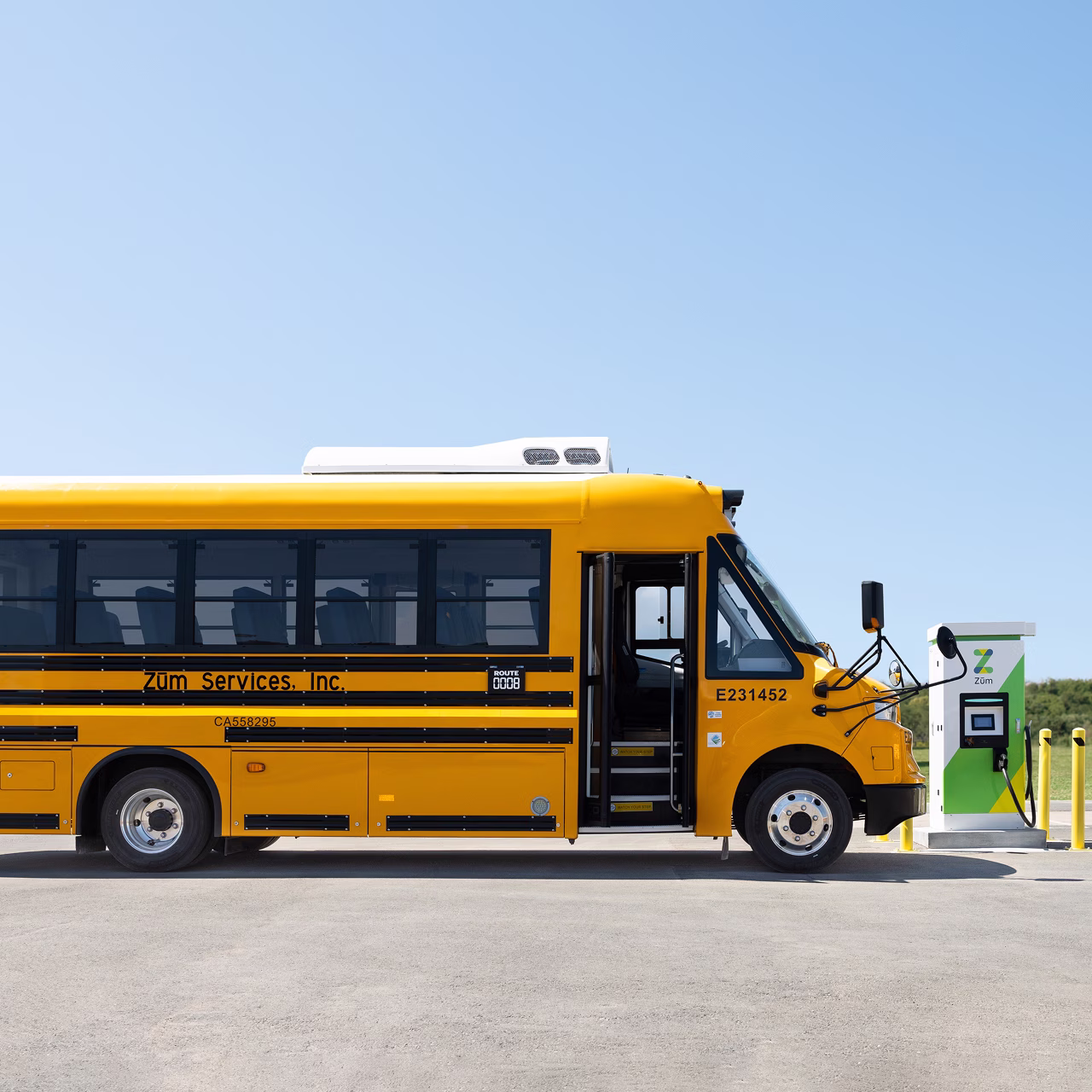 Side view of a Zum Services, Inc. electric school bus with its door open, parked next to a charging station.