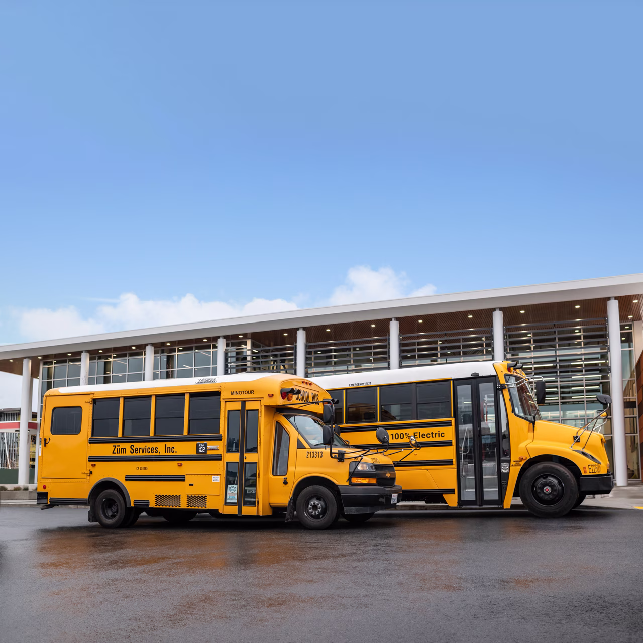 Two Zum Services, Inc. school buses, including a 100% electric model, parked outside a modern school building.