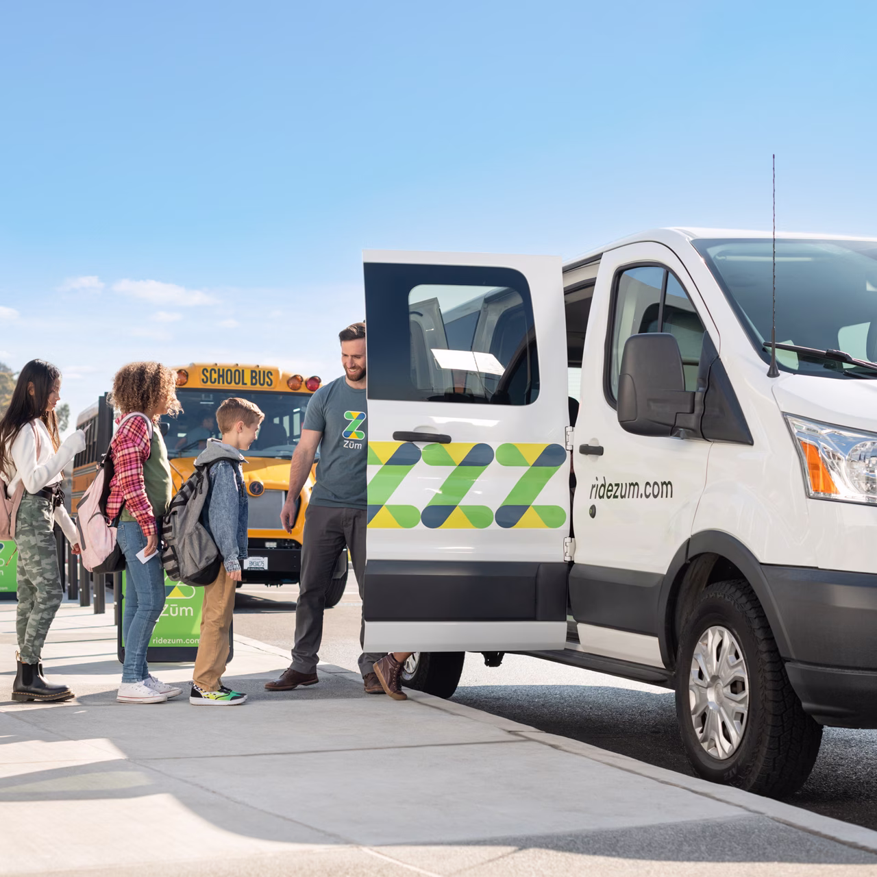 A Zum Services, Inc. driver welcoming a group of students into a branded Zum van, with a school bus parked nearby.