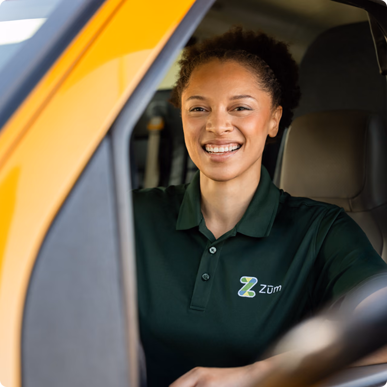 A smiling Zum Services, Inc. driver seated behind the wheel of a school bus.