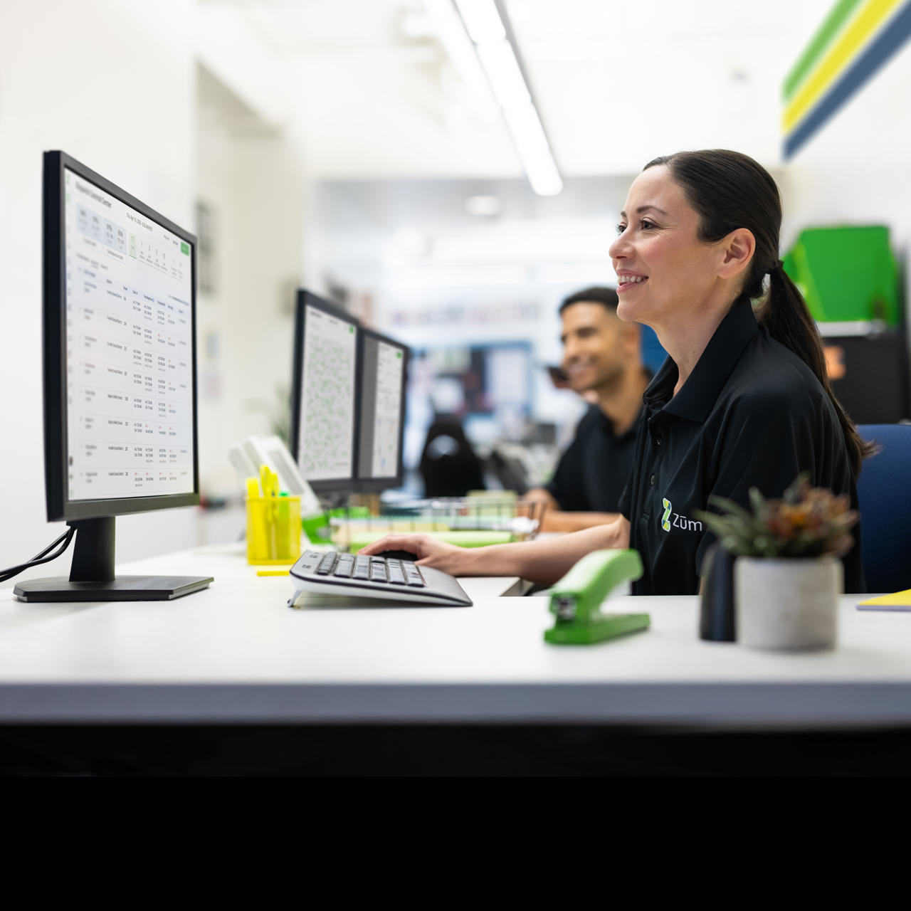 A smiling Zum Services, Inc. employee working at a dispatch desk with route data displayed on multiple monitors.