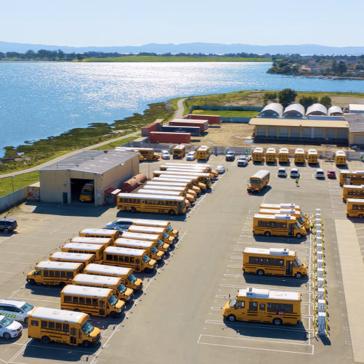 Aerial view of a Zum Services, Inc. bus yard with rows of school buses parked beside a waterfront maintenance facility.