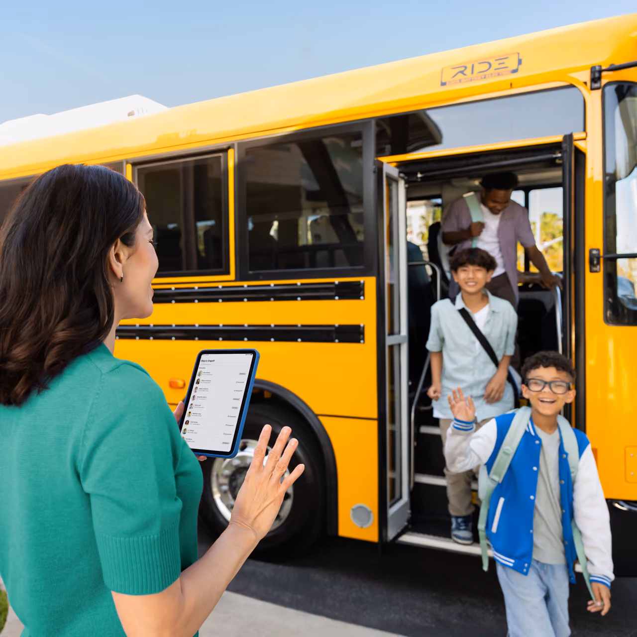 A staff member checking student pickup information on a tablet while students exit a Zum Services, Inc. school bus.