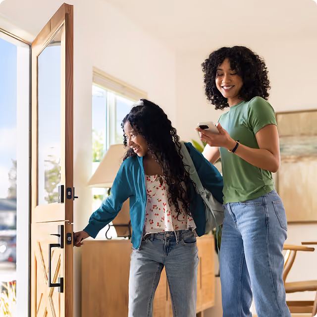 A child opens a front door while an adult smiles and holds a phone inside a bright home.