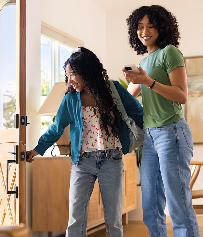 A mother checks the Zum app on her phone as her daughter heads out the door with a backpack, ready for her Zum ride to school.