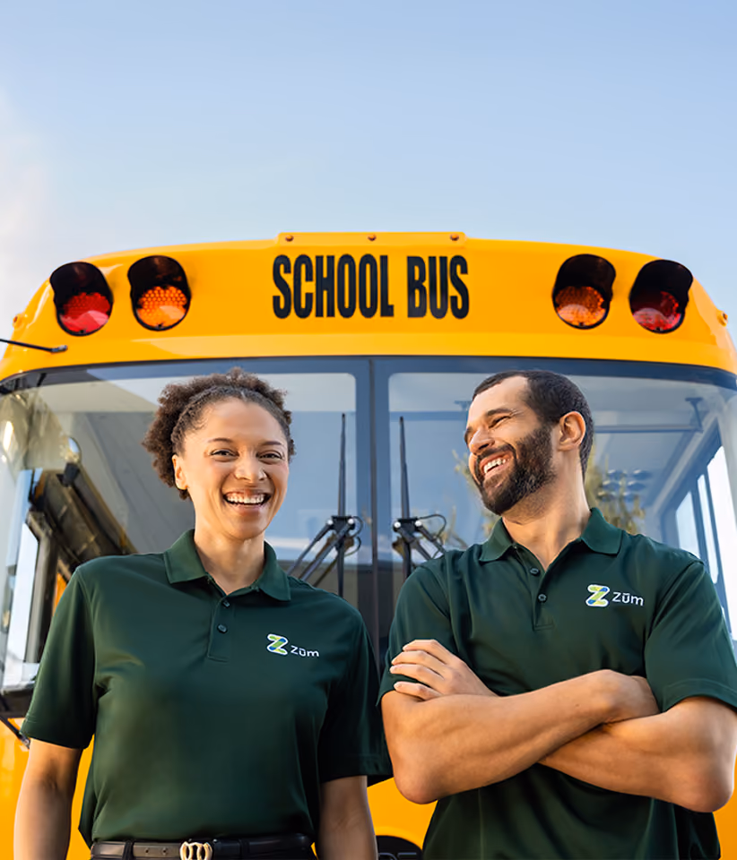 Two smiling Zum school bus drivers in green Zum polo shirts standing in front of their school bus.