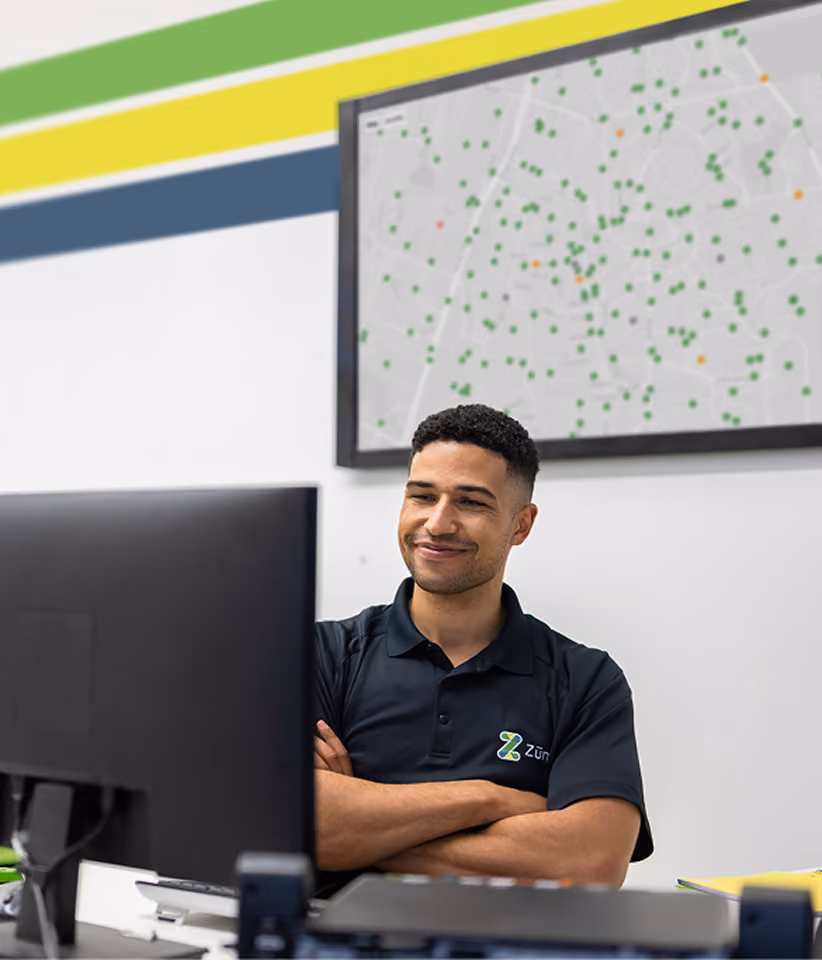 A smiling Zum operations team member at a yard office desk, with a live map of Zum vehicle locations displayed on the wall behind him.