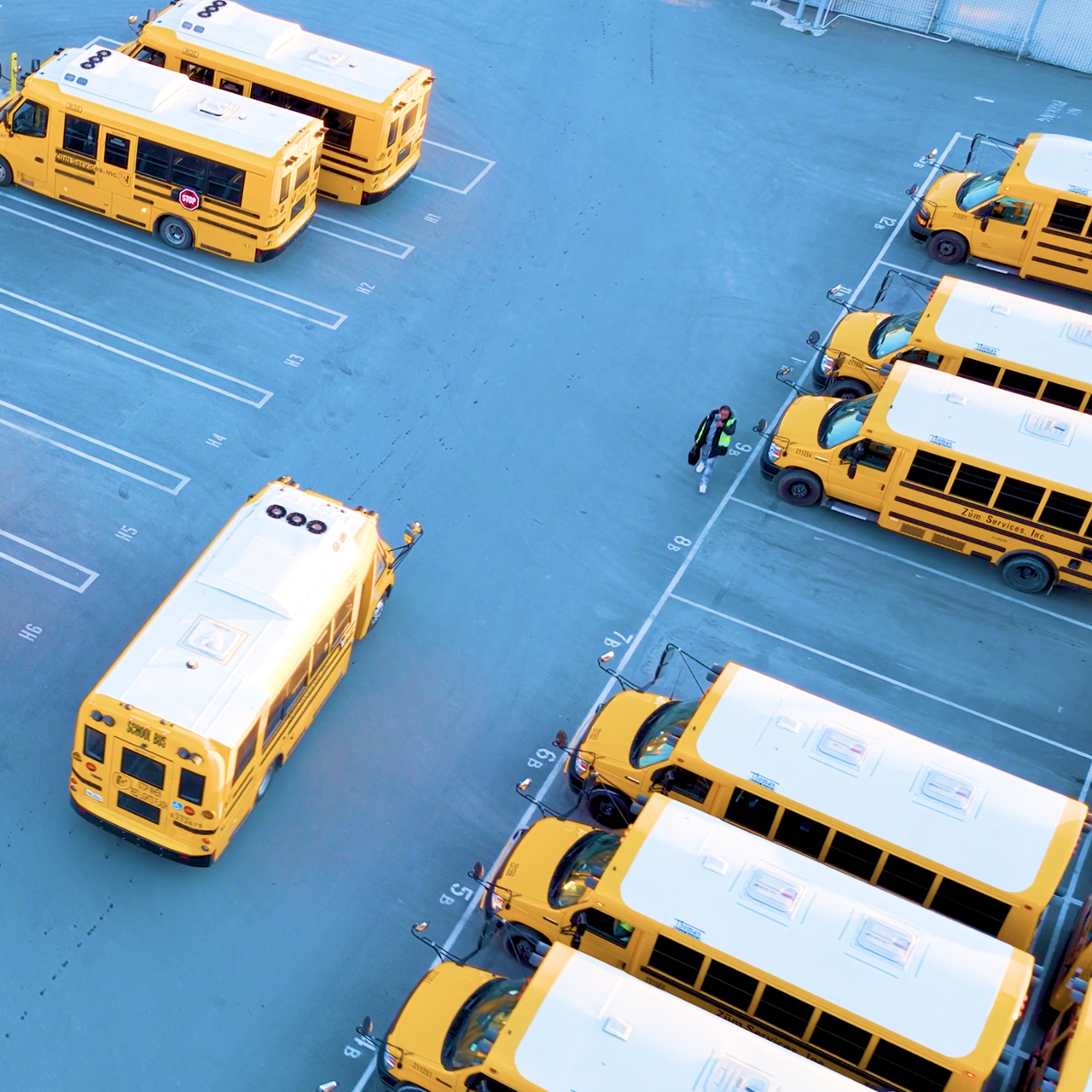 Aerial view of Zum school buses parked in numbered spaces at a bus yard, with a driver walking toward one of the buses.