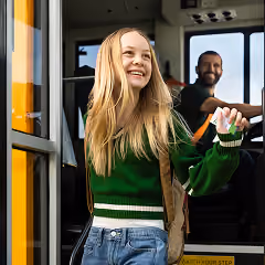 A smiling student exiting a Zum school bus, with the driver visible behind her.