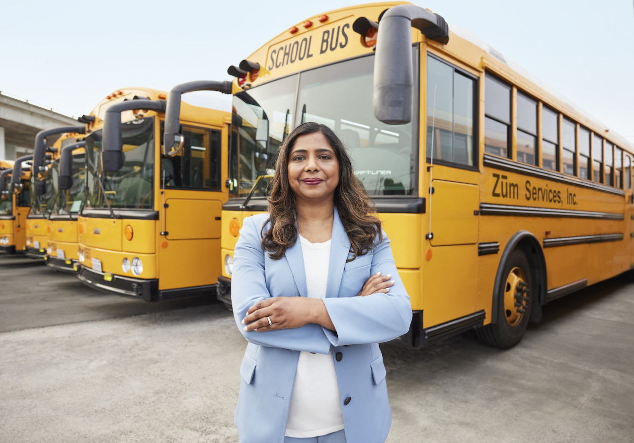 A woman in a light blue suit stands confidently in front of yellow school buses.