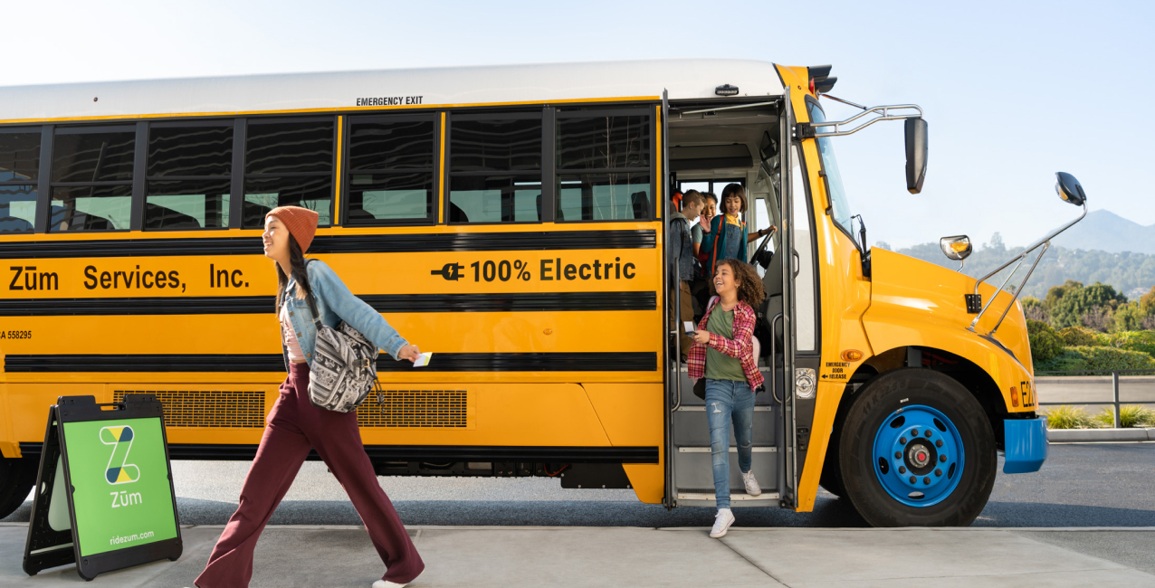 Students exit a Zum electric school bus while a girl walks past, smiling.