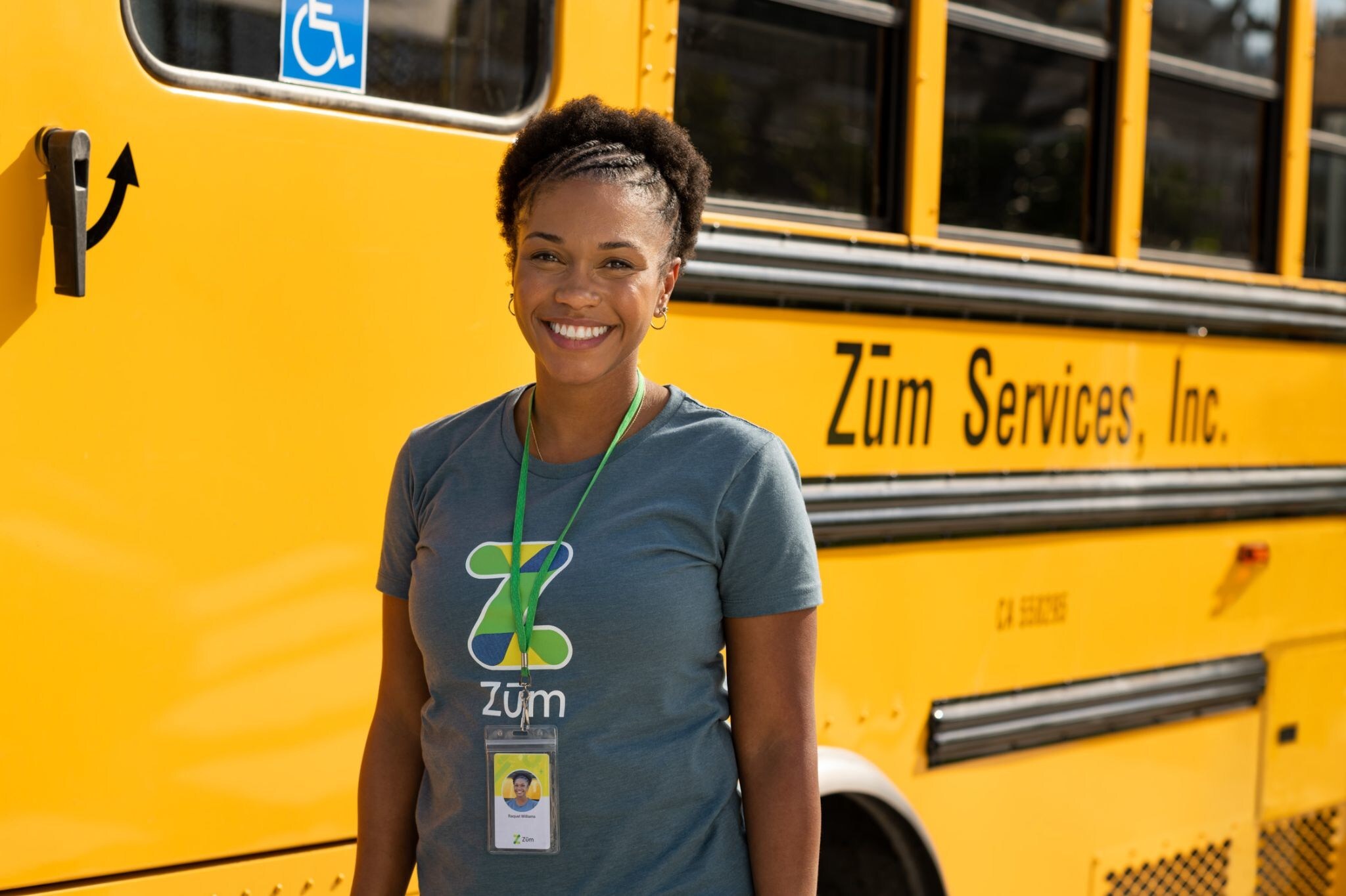 A Zum employee smiles in front of a yellow Zum services school bus, wearing a company shirt and ID.