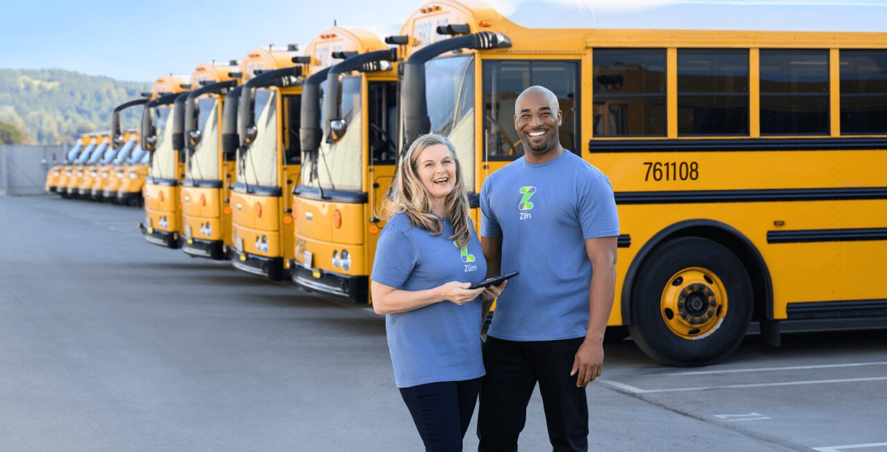 Two Zum employees stand in front of yellow school buses, smiling and holding a tablet.