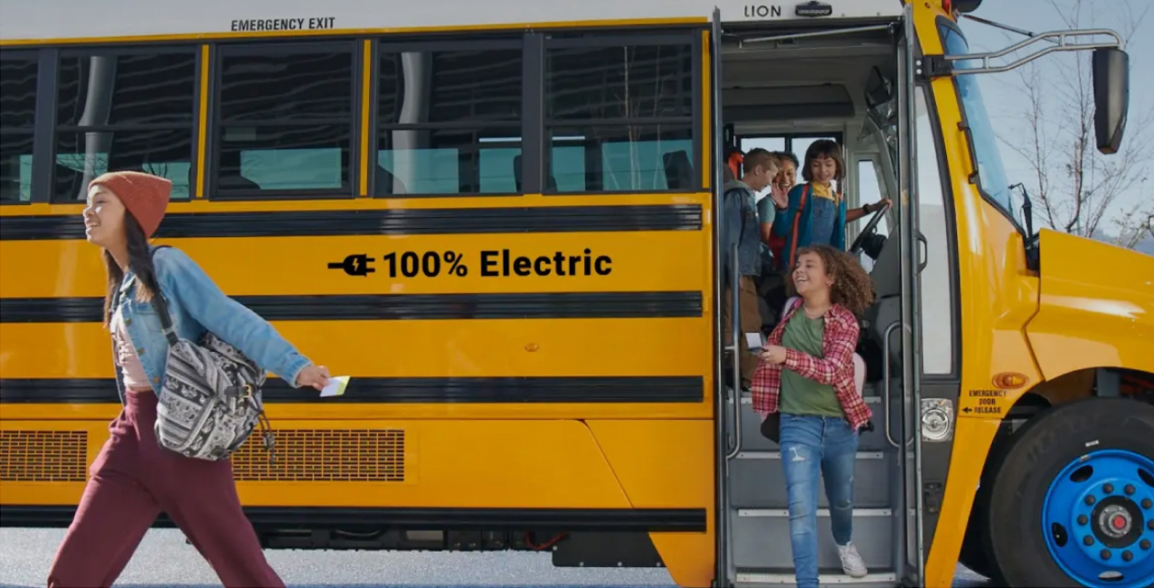 Students exit a yellow electric school bus labeled "100% Electric" in a bright atmosphere.