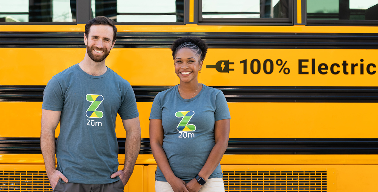 Two Zum team members stand smiling in front of a yellow electric school bus, showcasing the logo.