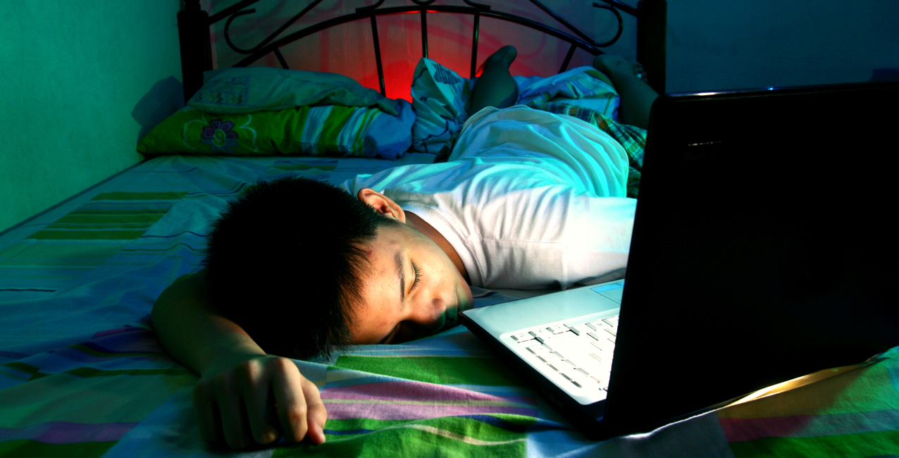 Child lying on bed with head on laptop, surrounded by colorful bedding, in dimly lit room.