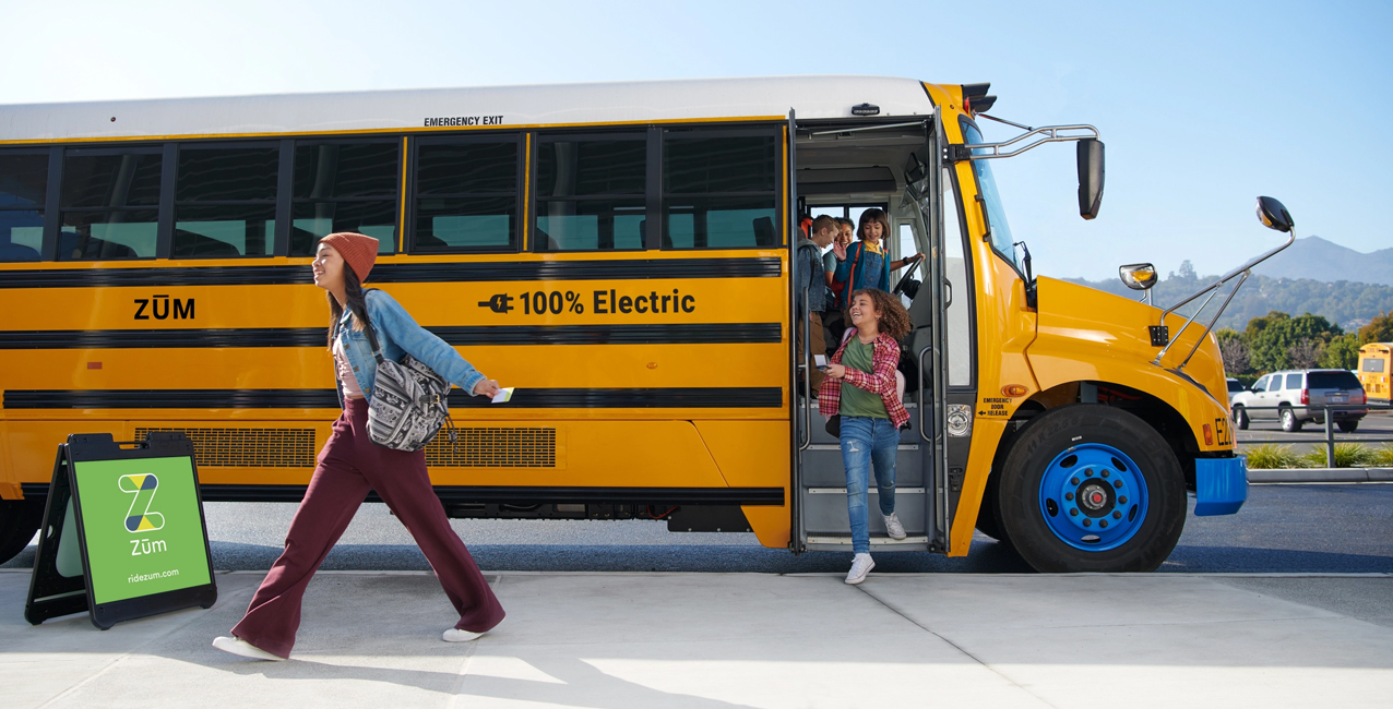 Students exit a yellow electric bus as a woman with a backpack walks by, near a Zum sign.