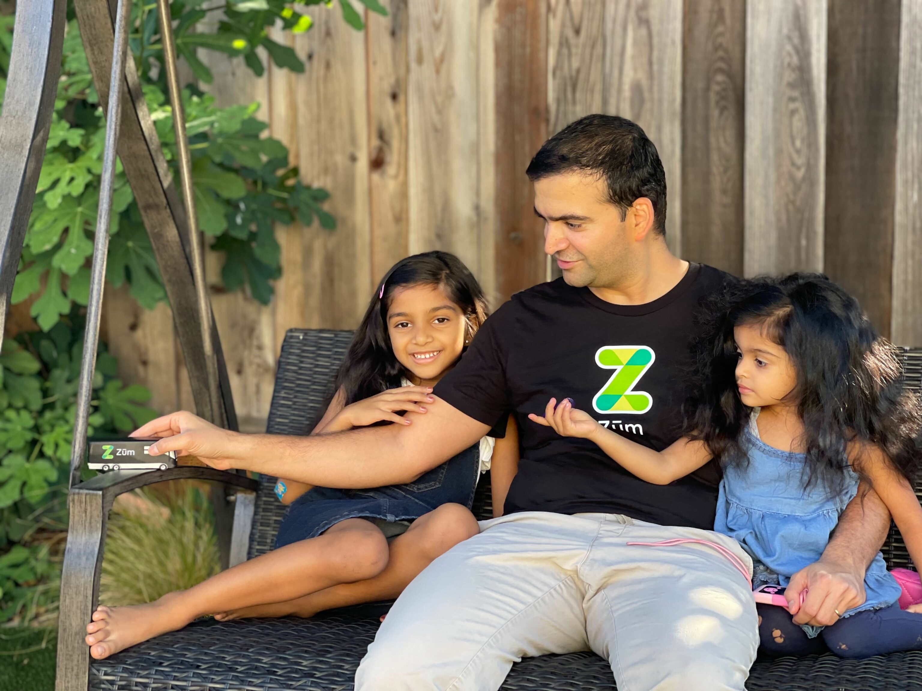 A man sits on a swing with two young girls, holding a Zum card, greenery in the background.